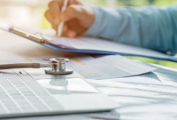 Physician taking notes next to a laptop with a stethoscope on top of it.