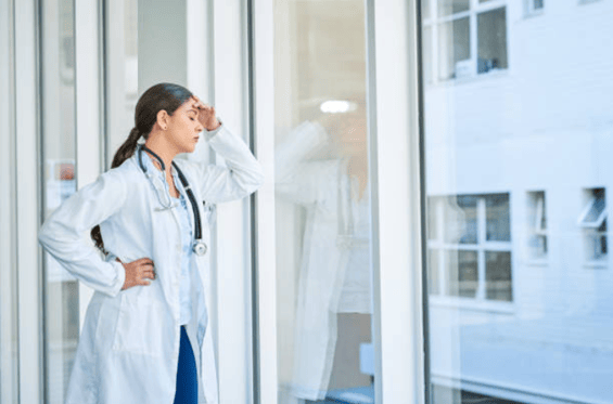 Brunette female doctor leaning against the window with her left hand on her forehead looking stressed