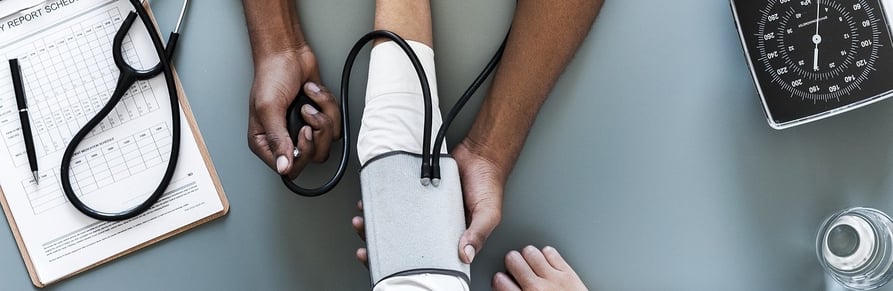 Blood pressure is being measured with a cuff and stethoscope; a clipboard and analog gauge are visible nearby.